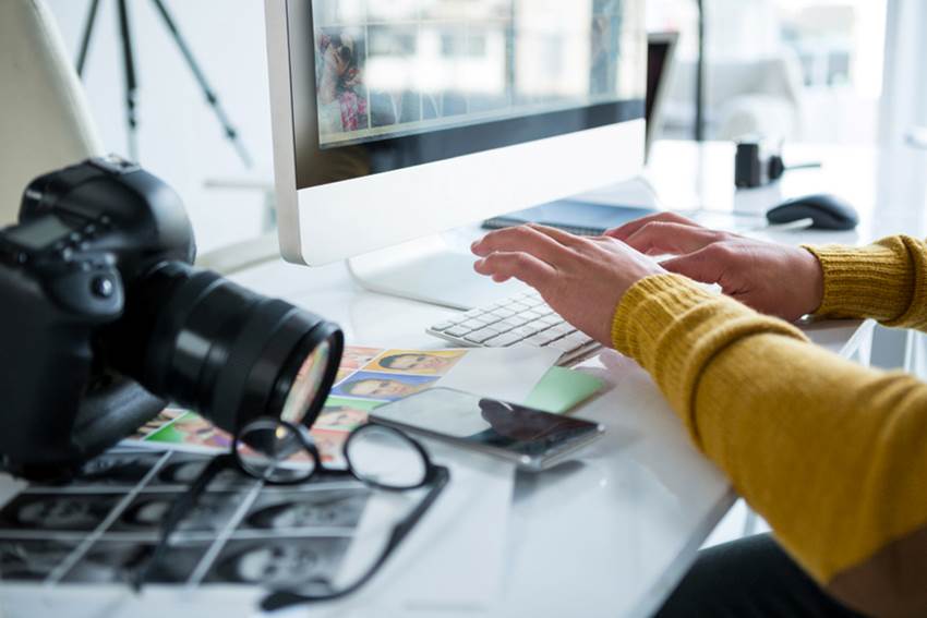 Fotógrafa fazendo o marketing digital da sua pequena empresa pelo computador com máquina fotográfica, óculos, celular e algumas fotos ao lado dela