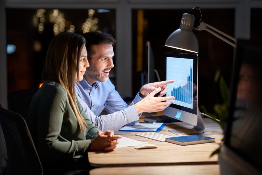 Duas pessoas analisando gráfico de barras crescente em um computador, discutindo formas de melhorar o custo de aquisição de cliente (CAC).