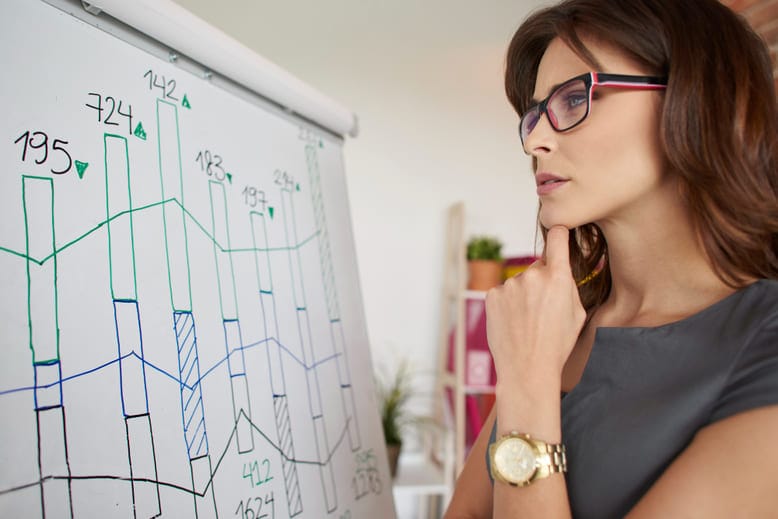Pessoa analisando gráficos estatísticos em um quadro branco, observando variações e tendências em dados numéricos.